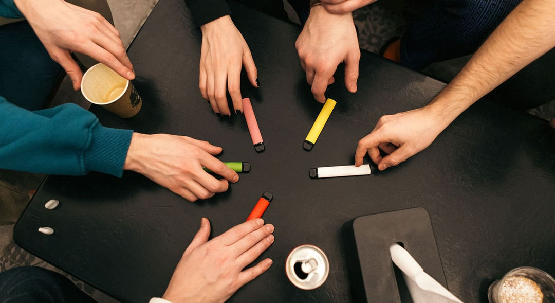Collection of disposable vapes on a table with people reaching for each one. Banner image for disposable vapes and nic salt e-liquid alternatives.