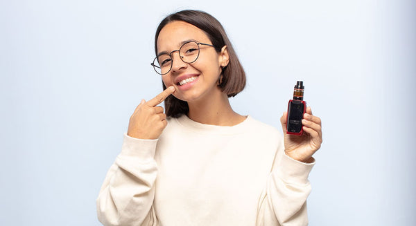 Woman holding vape device in left hand and pointing at her teeth with her right hand. Stood in front of a gradient white background.