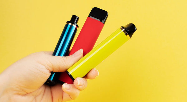 Person holding three different big puff vapes in their hand against a bright yellow background.